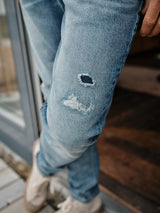 A person wearing Blue de Gênes Vinci Super Used Jeans - Used, with a torn knee and beige sneakers, stands by a glass door. The expertly crafted in Italy light blue jeans feature comfort stretch denim that is slightly faded for a casual, worn look.
