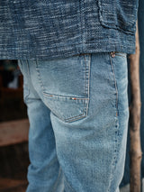 A person is wearing light blue denim jeans and a Blue de Gênes Kojima Jacket in navy, reminiscent of Japanese workwear. The focus is on the back pocket of the jeans with part of the jacket visible, set against a blurred wooden background.