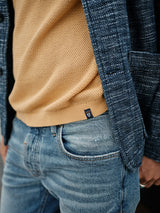 A person models the textured navy blazer over a Yellow Grain Filo knit polo sweater by Blue de Gênes, paired with faded blue jeans. The right hand casually rests in their pocket, seamlessly mixing casual and polished elements as the blazer drapes over the sweater.