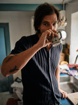 In a casual, slightly cluttered kitchen, a person with long hair sips from a patterned mug. Theyre dressed in the Blue de Gênes Compra Open Polo Knit in Dark Navy, featuring corozo nut buttons and retro stitch details, their tattoos adding to the unique aesthetic.
