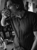 A man with medium-length hair drinks from a mug in a dimly lit kitchen. He wears the Compra Open Polo Knit in Dark Navy by Blue de Gênes, featuring corozo nut buttons. A tattoo decorates his left forearm as various kitchen items enrich the black and white image.