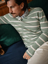 On a couch with dark green and blue cushions, a mustached man in Blue de Gênes Monte Stripe Knit - Green Bay and white pants wears a patterned scarf. He gazes sideways, casually resting one arm on the couch.