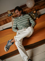 A man with a mustache sits on an orange couch, wearing a green striped sweater, Giacomo Bull WTH - White Denim jeans by Blue de Gênes, and blue sneakers. The scene includes wooden accents, a patterned pillow, and a round lamp. These Italian-crafted jeans offer timeless style.