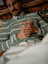 A person with a mustache is relaxing on a couch wearing a striped shirt and scarf, with rings including one turquoise stone washed ring. They are set against wooden walls and patterned cushions reflecting Italian craftsmanship. Theyre also sporting Blue de Gênes Giacomo Bull WTH - White Denim jeans.