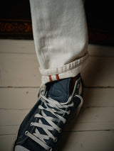 A close-up displays someone wearing Blue de Gênes Giacomo Bull WTH - White Denim with rolled white cuffs and red stitching, paired with blue canvas sneakers featuring white laces on a pristine white wooden floor.
