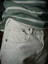 Close-up of a person wearing light grey, stone-washed Giacomo Bull WTH by Blue de Gênes, paired with a green and white striped shirt. The focus is on the Italian-made jeans front pocket, featuring a small metal emblem.
