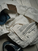 A pair of Blue de Gênes Giacomo Bull WTH - White Denim jeans, featuring a brown leather patch and text, is displayed on a wooden floor. This Italian-crafted stone-washed piece rests over a denim jacket with navy blue sneakers beneath.