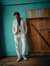 A man in the Blue de Gênes Leoni Desk Blazer - Natural stands relaxed against a turquoise wall and rustic wooden door, ready for summer parties, looking down with his hands in his pockets.