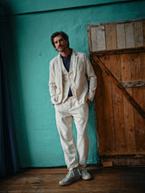 Wearing a Blue de Gênes Pantella Desk Gilet in natural, a person leans casually against a rustic wooden door. Complementing their look with Corozo buttons, navy shirt, and light gray sneakers on a wooden floor, the teal-painted wall adds charm to their effortless style.