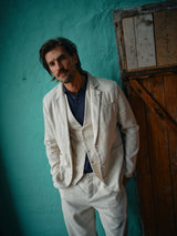 A person with medium-length hair and a mustache is wearing a cream-colored suit, a dark blue shirt, and Blue de Gênes Palto Desk Chino - Natural. Theyre standing against a teal wall with a wooden door, hands in pockets, looking at the camera.