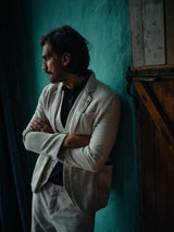 A long-haired, bearded man stands with arms crossed, wearing the Blue de Gênes Leoni Desk Blazer - Natural made from organic cotton, paired with a dark shirt. He leans against a teal wall in dim lighting, exuding effortless summer party charm.