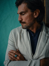 A man with dark hair and mustache dons the Leoni Desk Blazer - Natural by Blue de Gênes, crafted in an organic cotton-linen blend. He stands against a teal wall with arms crossed, featuring a turquoise ring on one hand, while gazing downward thoughtfully.