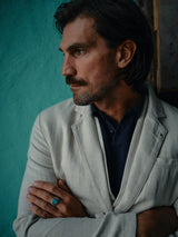 A man with long hair and a mustache stands with arms crossed, gazing to the side. He wears the Blue de Gênes Pantella Desk Gilet in Natural, adorned with Corozo buttons over a dark shirt, accentuated by a turquoise ring against a textured teal wall that contrasts beautifully.