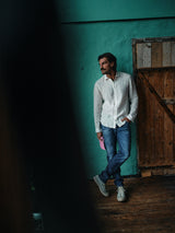 A man in a white shirt and Vinci Denova Jeans by Blue de Gênes leans casually against a wooden door, teal wall behind him. Holding a pink hat and looking aside, he wears white sneakers; the expertly crafted Italian denim ensures both ease and style on the wooden floor.