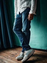 A person in a white shirt and Blue de Gênes Vinci Denova Jeans (Dark Blue Denim) stands against a teal wall with legs crossed. They wear white high-top sneakers and a silver ring; the rustic wooden floor and a partially visible dark curtain complete the scene.