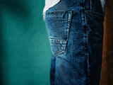 Close-up of a person wearing Blue de Gênes Vinci Denova Jeans in dark blue denim, showcasing the expertly crafted back pocket. The comfort stretch fabric enhances the relaxed vibe against a softly blurred teal background.