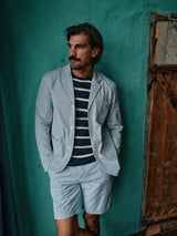 The mustached man wears a Blue de Gênes light blue blazer in fine line cotton, Elak Riga Shorts - Blue Stripe with Corozo buttons, and a navy striped shirt. He stands facing sideways against a textured teal wall and rustic wooden door, hands in pockets.