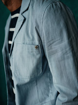 A person wearing the Blue de Gênes Gabbiano Riga Blazer - Blue Stripe, featuring a pocket and small cross pin, is seen from the side. The relaxed fit matches the dark teal background beautifully.