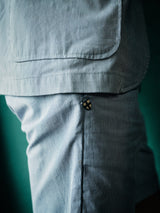 A close-up features a person in the Gabbiano Riga Blazer - Blue Stripe by Blue de Gênes, highlighting a pocket and a small stylized scissors logo. The light blue and white cotton yarn-dye stripes in a relaxed fit beautifully contrast with the dark green background.