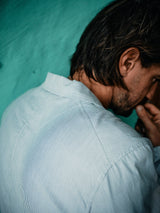 A man with brown hair, seen from behind and in profile, wears the Blue de Gênes Gabbiano Riga Blazer in blue stripe. He stands against a teal wall, hand near his face and head slightly turned right.