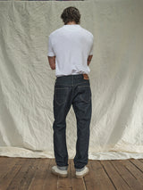 A person with shoulder-length hair, in a white t-shirt, Giacomo Selvedge Jeans - Raw Denim by Blue de Gênes, and light sneakers stands facing away from the camera. The backdrop is textured off-white fabric, and the floor looks like wood.