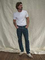 A man with medium-length hair, wearing a white t-shirt, Blue de Gênes Vinci Topaz Jeans - Rinse Wash, and white sneakers stands with hands in pockets against a wrinkled white fabric backdrop. A wooden floor adds a rustic touch.