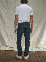 A person with shoulder-length hair stands with their back to the camera, dressed in a white T-shirt, cream sneakers, and Vinci Topaz Jeans - Rinse Wash by Blue de Gênes. The setting features a beige fabric curtain and a wooden floor.