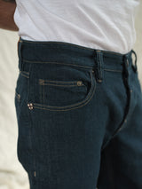 A close-up highlights the Vinci Topaz Jeans - Rinse Wash by Blue de Gênes, crafted in Italy from Comfort Stretch Denim. The dark blue jeans feature visible stitching and a small checkered flag tag near the front pocket, paired with a crisp white T-shirt against a blurred, neutral-toned backdrop.