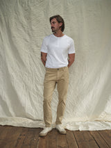 A man with medium-length hair and a mustache stands on a wooden floor in a vintage look. He wears a white henley shirt with Blue de Gênes Menzo Dobby Chino - Khaki pants, featuring comfort stretch. His hands are behind his back against a wrinkled white fabric backdrop.