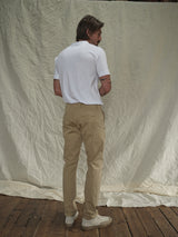 A man stands sideways on the wooden floor, wearing a white T-shirt with Menzo Dobby Chino - Khaki pants by Blue de Gênes. He presents a vintage look against a backdrop of wrinkled off-white fabric, showcasing comfort stretch for effortless style.
