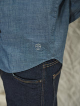 Close-up of a person wearing the Sandro Chambray Shirt by Blue de Gênes, featuring an embroidered shield logo, paired with dark blue jeans against a soft, light-colored fabric background.