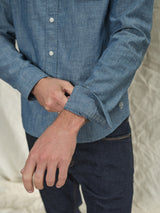 A person in a Sandro Chambray Shirt by Blue de Gênes is adjusting their sleeve while wearing dark blue jeans against a light, crumpled fabric background.