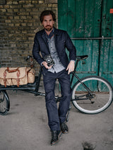 A man with a beard and styled hair wears the Blue de Gênes Amos Blazer in dark navy, paired with a striped shirt, scarf, and boots. He stands by a bicycle with a tan bag, holding a vintage camera before green doors and an aged brick wall.