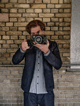 Wearing the Blue de Gênes Amos Blazer in Dark Navy over a striped shirt, someone stands by a brick wall and lifts a vintage camera to their face as if snapping a photo.
