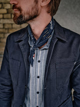 A bearded man wears the Blue de Gênes Amos Blazer in dark navy over a blue striped shirt and a blue polka dot bandana, with a blurred brick wall in the background.