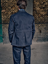 A person with brown hair, wearing the Blue de Gênes Amos Blazer in Dark Navy, stands with their back to a weathered brick wall. The photo captures only the rear view of the individual.