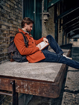 A bearded person in an orange coat and blue jeans sits on a concrete ledge by a brick wall, writing in a BDG Leather Journal - Brown. Beside them, a dark backpack made of premium bull leather enhances their vintage style in the rustic outdoor setting.