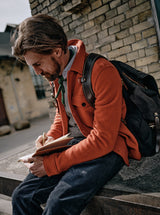 A man with short brown hair, a beard, and a vintage style in an orange jacket and green scarf sits on a stone bench. He writes thoughtfully in a BDG Leather Journal by Blue de Gênes, made of premium bull leather, set against brick buildings.
