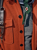 Close-up of an outfit featuring the Pampa Overshirt in Ochre Red by Blue de GĂȘnes with a pocket and Corozo buttons, layered over a gray boiled wool sweater. A green necktie with a white cross emblem contrasts nicely, while a dark leather strap with clasp crosses the shoulder.