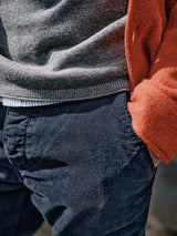 A person in a Blue de Gênes Monte AW Knit in Stormy Grey, layered over a striped shirt, stands with hands in the pockets of dark blue textured pants. An orange garment on the shoulders adds to the cozy and casual look.