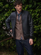 A man stands outdoors before lush foliage, wearing the Rufo Shearling Jacket - Scarab by Blue de Gênes over a brown cardigan, blue denim shirt, and dark jeans. He gazes thoughtfully to the side.