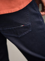 A close-up shows a person wearing Blue de Gênes Vinci Venom Blk Jeans - Dark Denim with a rust shirt. The focus is on the back pocket, featuring subtle stitching and a small embroidered red, white, and blue logo. Made in Italy.