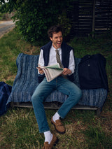 A man laughs outdoors on a blue blanket-covered bench, holding an open newspaper. He wears jeans, a white shirt, the Blue de Gênes Nekutaki Tie #E - Sand Pattern, a navy vest, brown shoes, and has a dark jacket beside him.