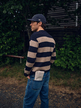 A man in a navy and beige striped sweater, blue cap, and Blue de Gênes Marco Used Jeans (Used) stands outdoors with his back turned. A newspaper peeks from his pocket as green bushes and a wooden wall form the backdrop.