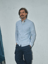 A person with a mustache stands against a plain white wall, wearing a light blue button-up shirt, a patterned scarf around the neck, and Blue de Gênes Mustang Cargo Pant in Navy. Hands rest casually in their pockets as they look away from the camera.