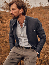 A bearded man in a Blue de Gênes Casella Officers Blazer - Dark Navy over a striped shirt and khaki pants sits on a fence, surrounded by dry autumnal foliage that enhances the rustic and natural ambiance.
