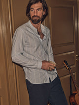 A bearded man stands in a room, holding a cup and wearing a Blue de Gênes Basso Hackney Shirt - Blue Stripe, paired with dark pants. He leans against a brown door with an acoustic guitar nearby. The lighting is warm and subdued.