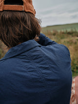 Wearing a Gervaso Tarlo Blazer in navy by Blue de Gênes and a brown cap with corozo buttons, a person stands back to the camera, gazing over grass. Brown hair peeks from the cap as they survey the landscape under an overcast sky.