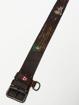 The Gari Belt by Blue de Gênes stands out with its metal buckle and ornate details, including a shield emblem, script writing, and petite images like a green stamp and ship. Crafted from premium leather in brown, its an accessory that contrasts beautifully against a white background.