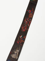 The Gari Belt - Brown by Blue de Gênes is a long, narrow fabric strip with intricate, colorful embroidery of people in traditional clothing, flags, and symbols against a dark background that highlights its vivid detail.